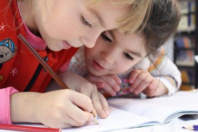 Two young girls sitting at a table, writing in a book together.